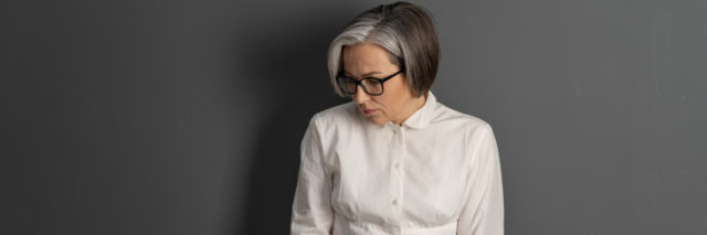 A white middle-aged woman with gray hair and glasses looking down, sad, against a black wall