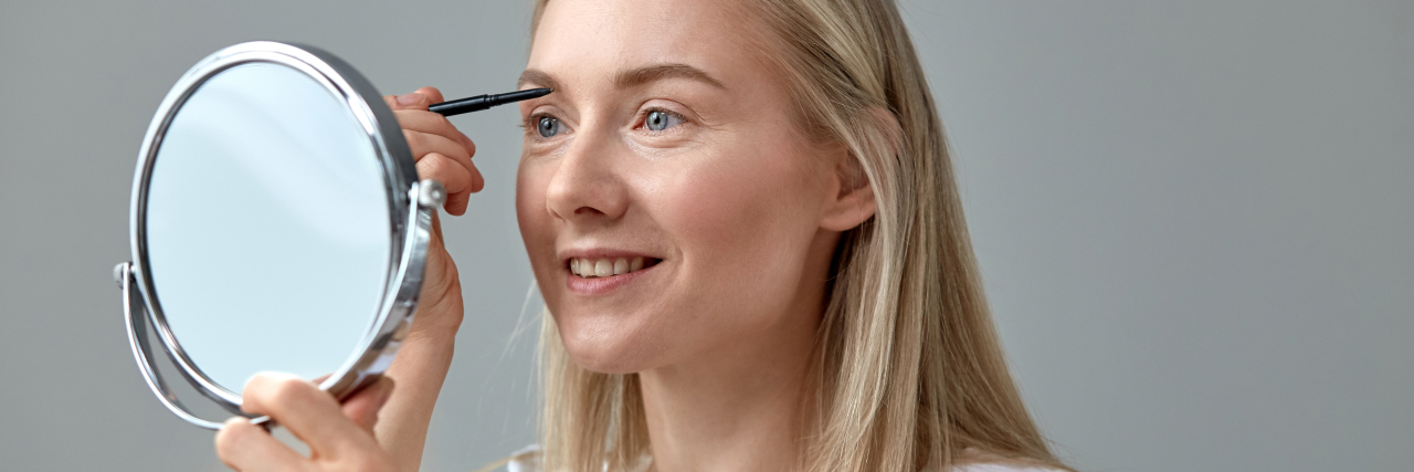 8 Makeup and Beauty Tips for Women With Chronic Illnesses Woman putting on makeup in front of a mirror.