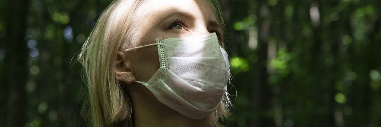 How I Cope With Pandemic Stress as an Immunocompromised Person A woman facing the sun in a dark forest wearing a coronavirus mask during global pandemic.