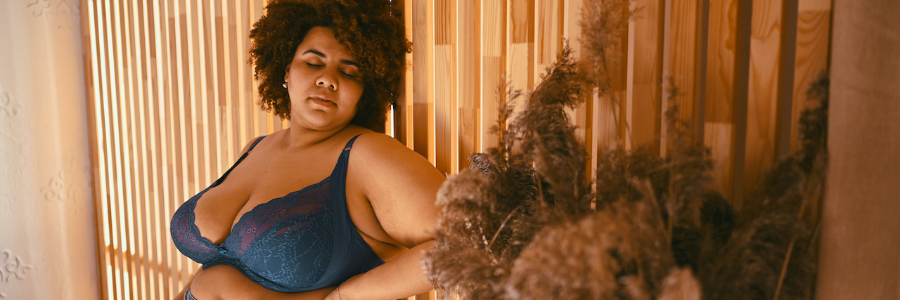 A Black woman in jeans and a blue lace bra, leaning against a wall.