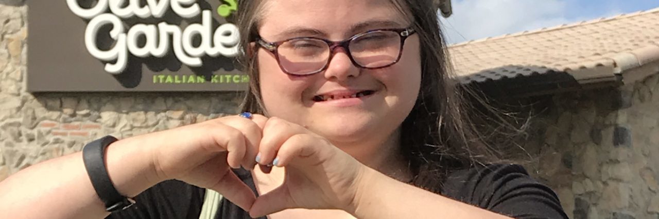 Going Beyond Down Syndrome Awareness and Promoting Employment Rachel outside Olive Garden making a heart sign with her hands.