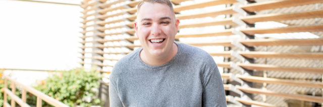photo of the contributor, Kyle Elliott, posing and smiling with wooden blinds or structure behind him and lots of light