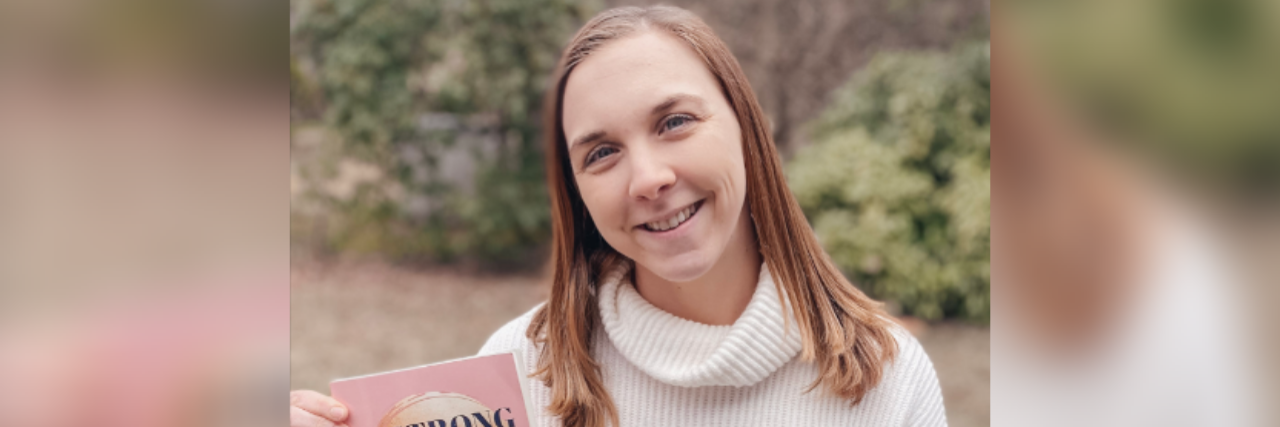 Photo of author Kelsey Buckholtz, a young white woman with ginger hair smiling outside holding her book