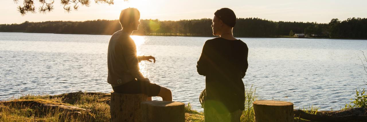 4 Things Men Can Do to Manage Their Anxiety Two young men sitting on tree stumps talking with a lake in the background and sun shining