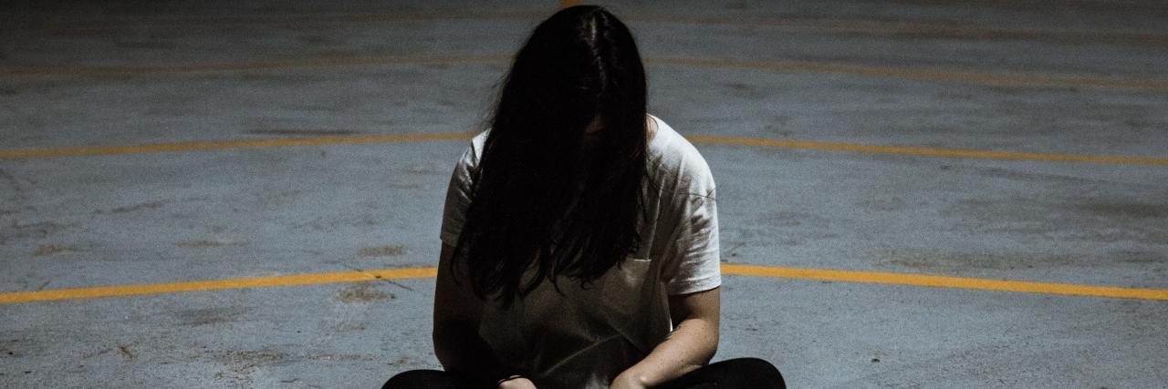 A young white woman sitting on a road with her head down, hair covering her face, sad