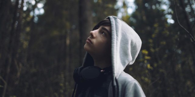 photo of a young teenage boy wearing a hooded sweatshirt and looking up at trees in a forest