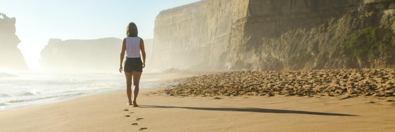 Mental Health Recovery Feels Like Two Steps Forward, One Step Back photo from behind of a woman walking along a beach, with the camera focused on her footprints