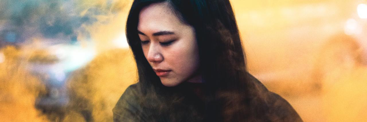 How Asian Hate Has Triggered My PTSD and Suicidal Thoughts photo of an Asian American woman standing in orange smoke, facing away from the camera with her eyes closed and downturnd