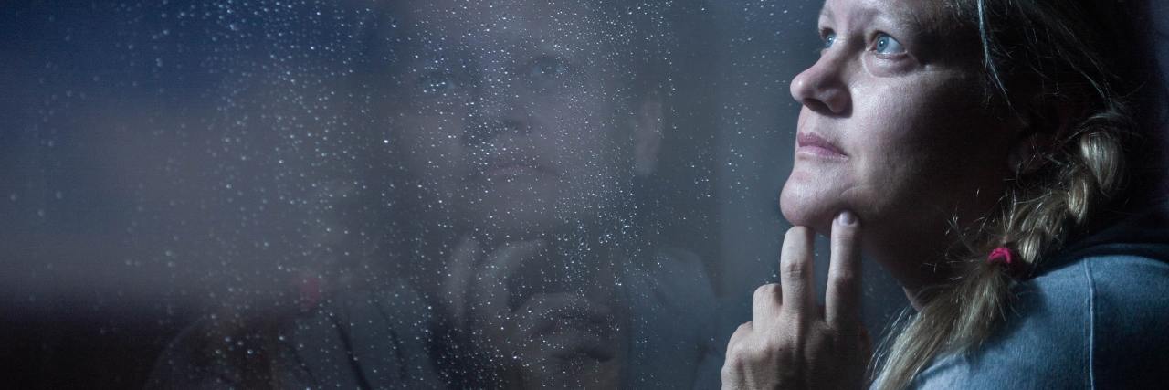 photo of a woman looking out of a rainy window at night with her reflection showing and face illuminated