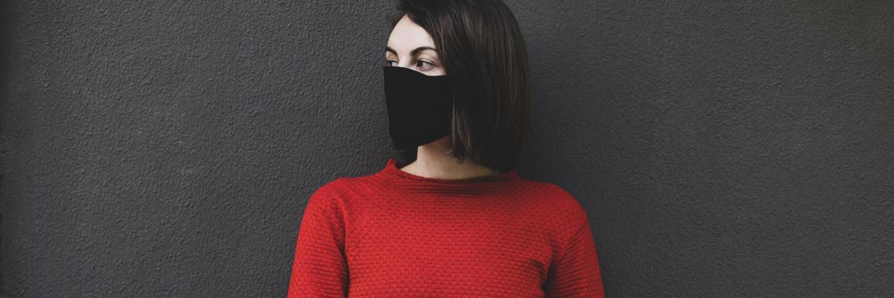 The Mental Health 'Echo Pandemic' That Will Come After COVID-19 photo of woman in a red top and black face mask, looking away and standing against a dark background