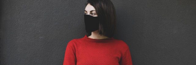 photo of woman in a red top and black face mask, looking away and standing against a dark background
