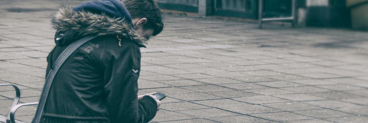 A Letter To the Bully Responsible for My Addiction and Depression photo of a young man sitting on a bench outside with a sachel, looking down at his phone