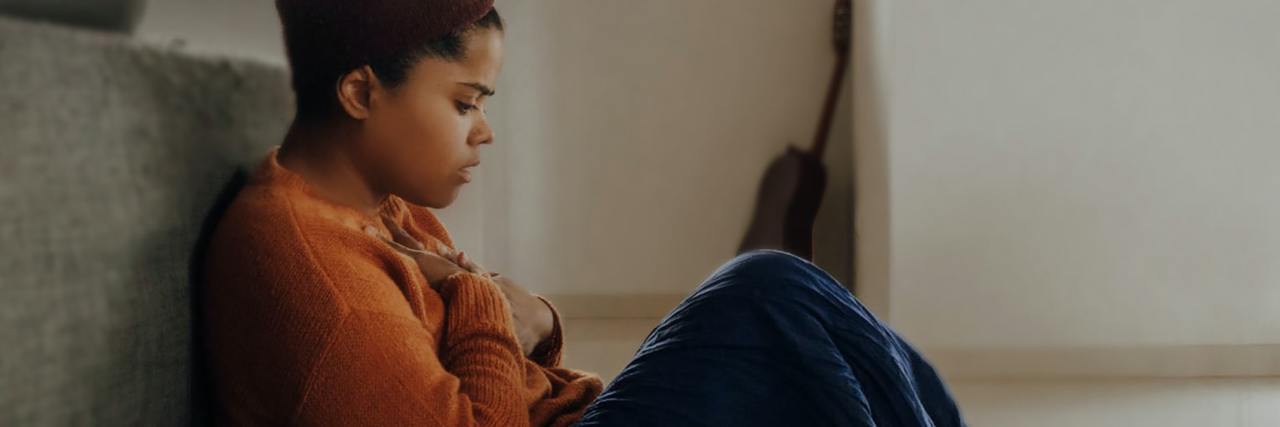 Therapy Techniques to Improve Your Mental Health and Well-Being photo of a woman sitting on the floor, looking anxious, with her hands to her chest and her back resting against a couch