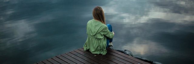 photo of a woman sitting on a wooden dock, hugging her knees, with the water reflecting the sky