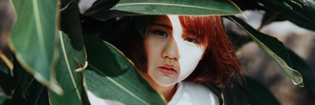 An Asian woman peaking out from behind large green leaves