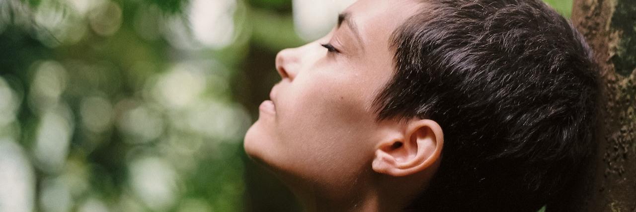 The Cycle of Trauma Caused by Insufficient Mental Health Resources A photo of a woman with short hair closing her eyes and leaning against a tree, looking up