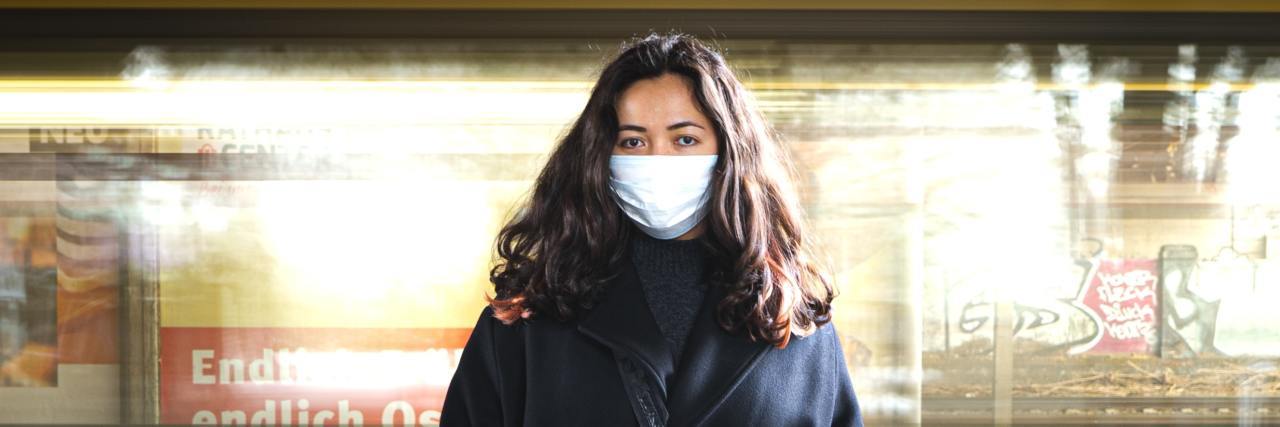 Dealing With COVID-19 Anxiety One Year Later photo of a woman wearing a face mask, standing looking into a camera with a train moving past behind her