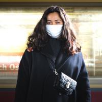 photo of a woman wearing a face mask, standing looking into a camera with a train moving past behind her