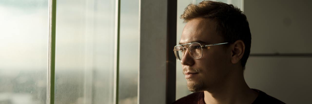 A young white man in a red sweater with glasses looking out a window not smiling