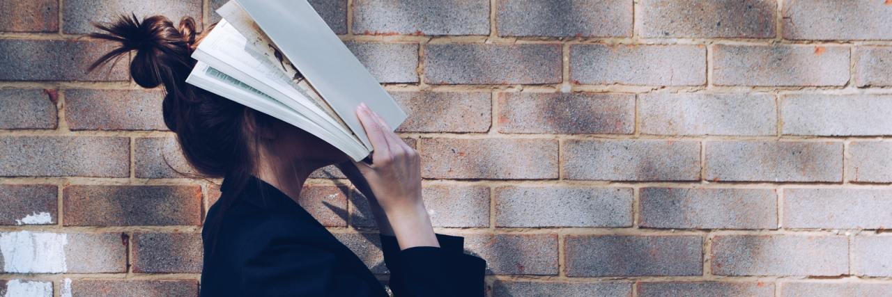 photo of a young woman holding a book to cover her face, standing in front of a brick wall
