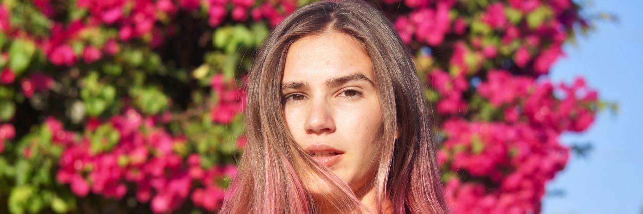 Life With Chronic Illness Has Taught Me to Appreciate Everything A young white woman with pink highlights in her hair standing in front of a tree looking off