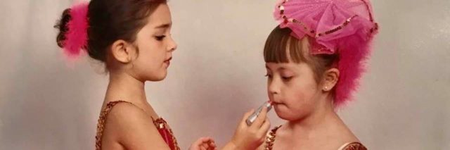 The author's daughter, Yazzy, in a dance class as a child. She's waring a pink dance costume, and another little girl is helping her put on makeup.