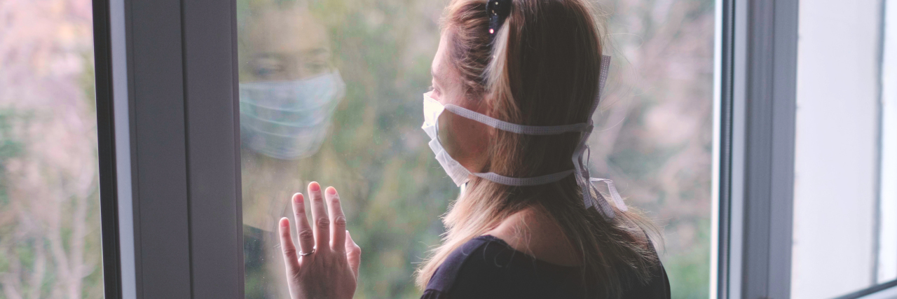 A young white woman with brown hair wearing a mask inside with a hand on the window