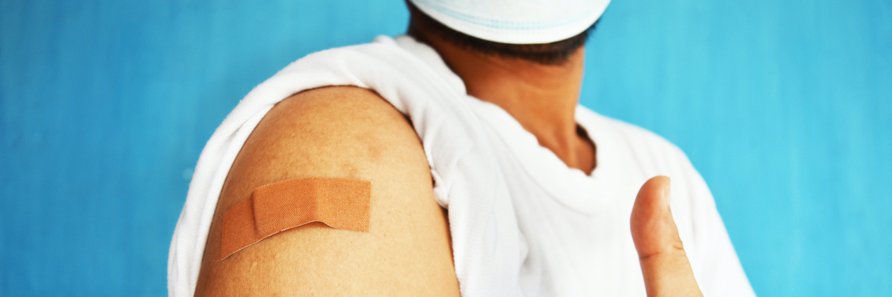 A Letter to Chronic Illness Patients With COVID-19 Vaccine Anxiety A man holding up a thumbs up with a bandage on his arm after getting the covid-19 vaccine