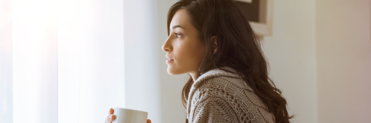 A young woman in a sweater sitting down looking out the window holding a cup of coffee