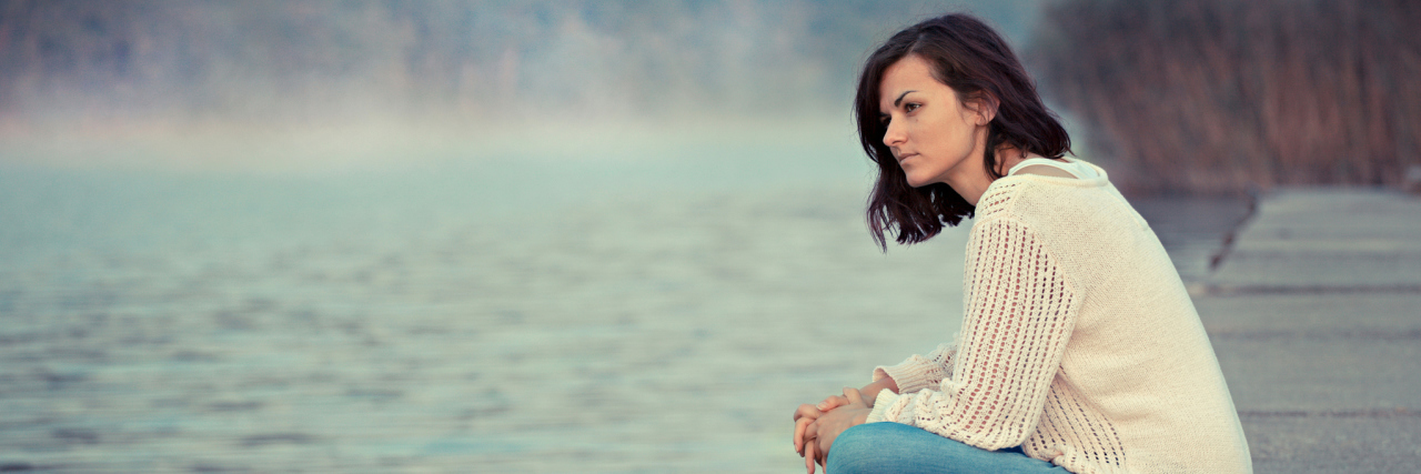 A young white woman sitting by a lake looking out