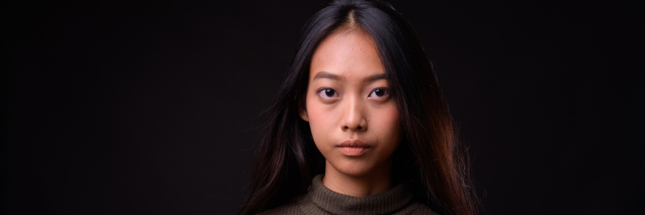 5 Unhelpful Things to Do or Say to Someone Who Self-Harms A young Asian woman with a serious face looking into the camera against a black background