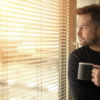 Young man standing by the window drinking a cup of coffee, looking outside