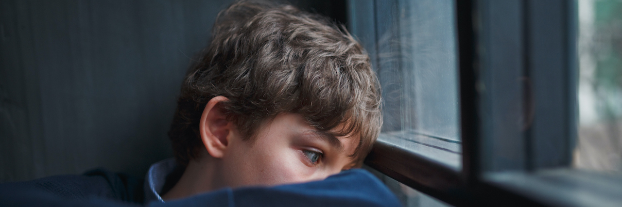 Pensive teenager with blue eyes in a blue shirt and jeans leaning against a window pane
