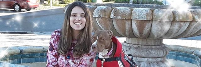 Jennica sitting by a fountain with her service dog, a brown pit bull.
