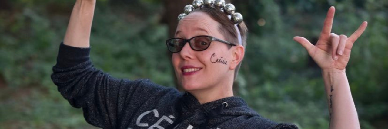 Read This If You Struggle With Reparenting Yourself in Trauma Therapy photo of the contributor giving horns signs with her hands, wearing sunglasses and a sweater which reads "Celine Dion Vegas." She is standing against a backdrop of trees.