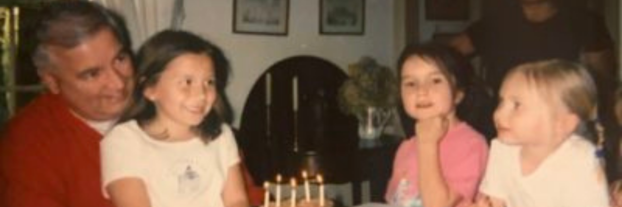 Dealing With Grief Not Even My Trauma Prepared Me for Photo of author sitting on her father's lap as a child in front of a birthday cake with her 2 sisters at the table