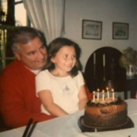 Photo of author sitting on her father's lap as a child in front of a birthday cake with her 2 sisters at the table