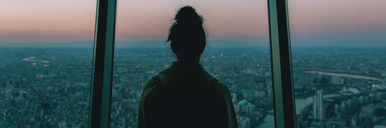 I'm Scared for My Mental Health in a World After COVID-19 A young female with her back to the camera looking out of a window at sunset