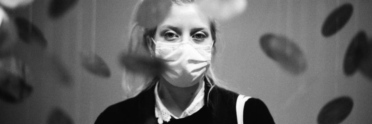 black and white photo of a woman wearing a face mask and looking into the camera, with disks suspended between the camera and her