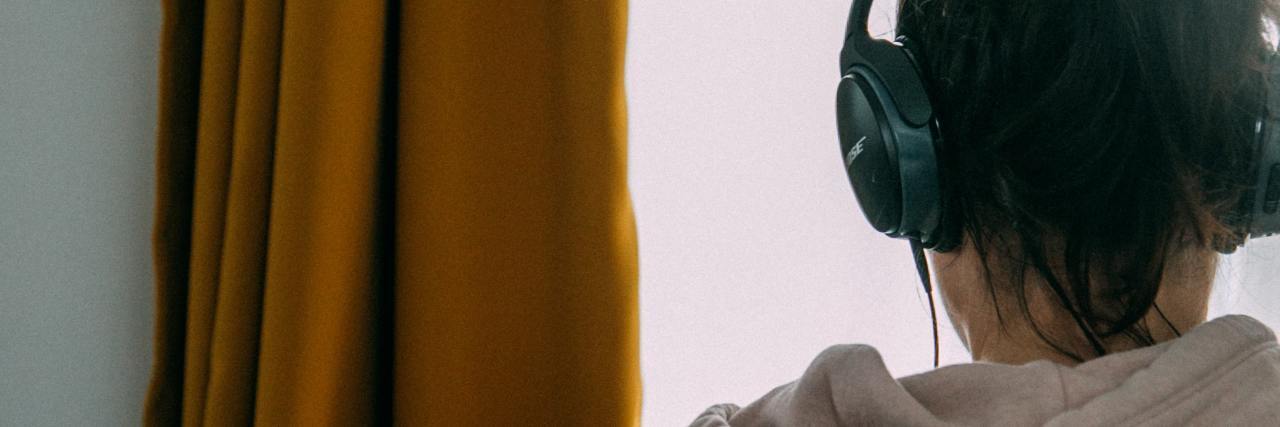 Why Listen to 'the Manic Episodes,' a Queer Mental Health Podcast photo taken from behind of a young woman in front of a window, listening to a podcast or music on headphones