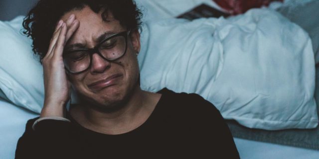 photo of a woman crying and upset while kneeling beside bed, her head in her hand