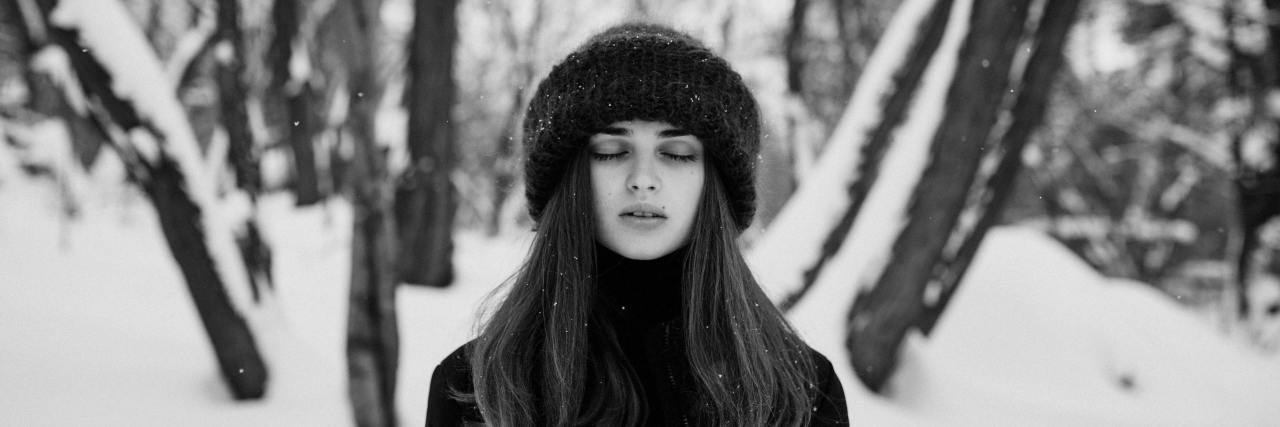 black and white photo of a woman standing in snowy woods with closed eyes, facing camera