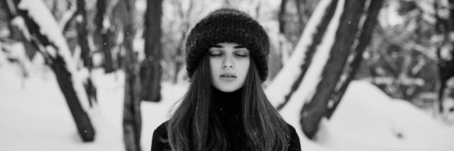 black and white photo of a woman standing in snowy woods with closed eyes, facing camera