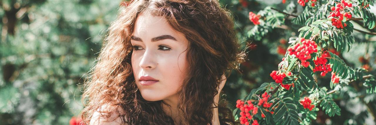 What I Want You to Know About My Fibromyalgia A young white woman with curly red hair standing in front of a bush looking away, serious facial expression