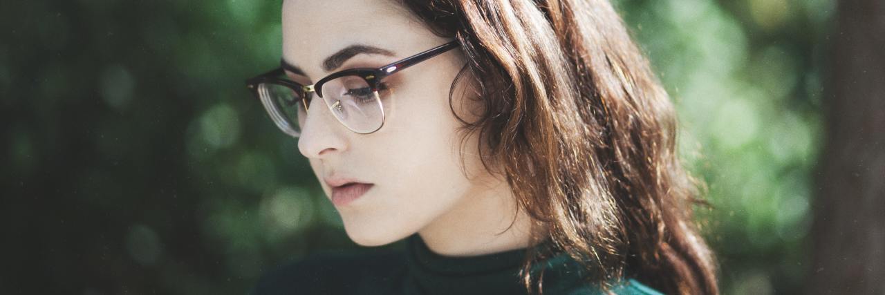 Should People Forgive Abusers After a Public Apology? photo of a young woman wearing glasses, looking down and away from camera with blurry foliage behind her