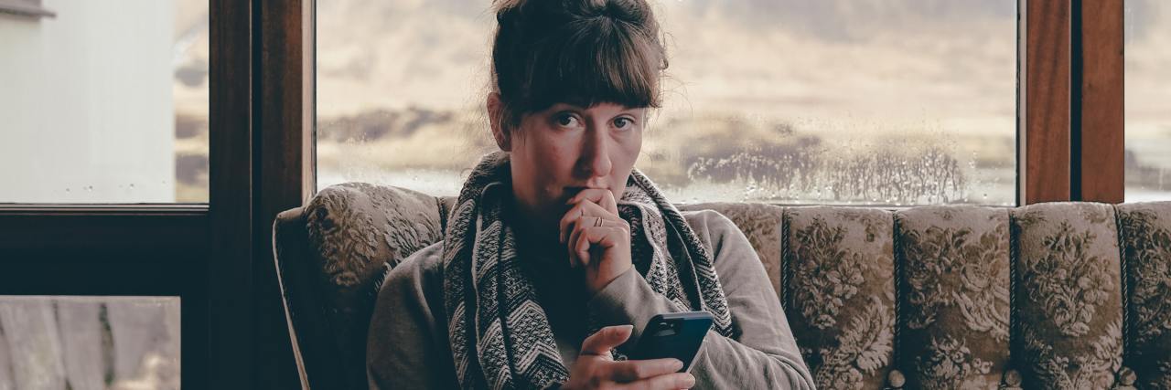Family Comparing Your Mental Illness With Someone Else's photo of a woman looking up into the camera from a sofa, phone in her hand, with a window behind her and her other hand raised to her mouth