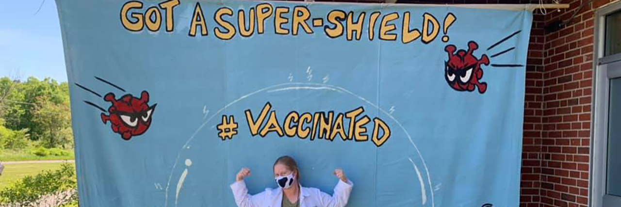 What to Say If Asked Why You're Vaccinated and Still Wearing a Mask Kateland wearing a mask in front of a Vaccinated banner with cartoon viruses being repelled by a bubble..