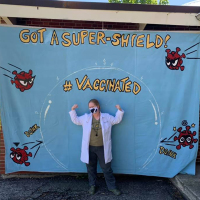 Kateland wearing a mask in front of a Vaccinated banner with cartoon viruses being repelled by a bubble..
