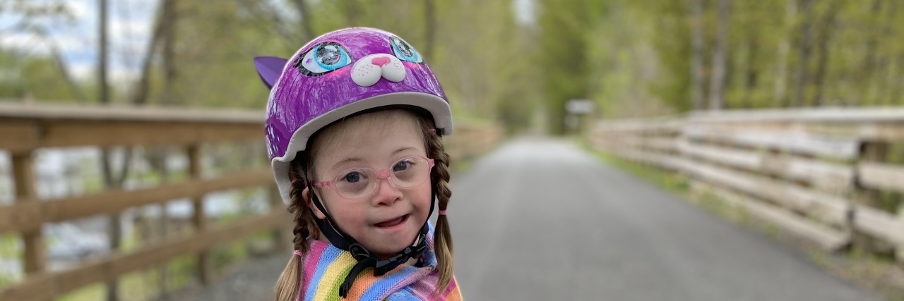 To My Daughter With Down Syndrome: You Belong Here Lily wearing a rainbow striped shirt and purple helmet.