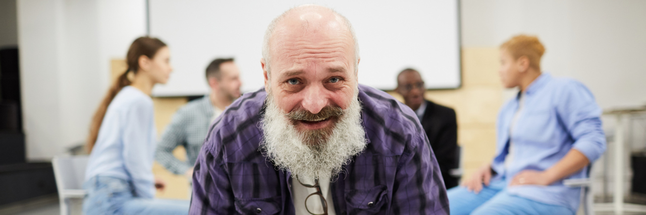 A bearded elderly white man in a flannel shirt sitting in a chair smiling with a support group behind him in a circle